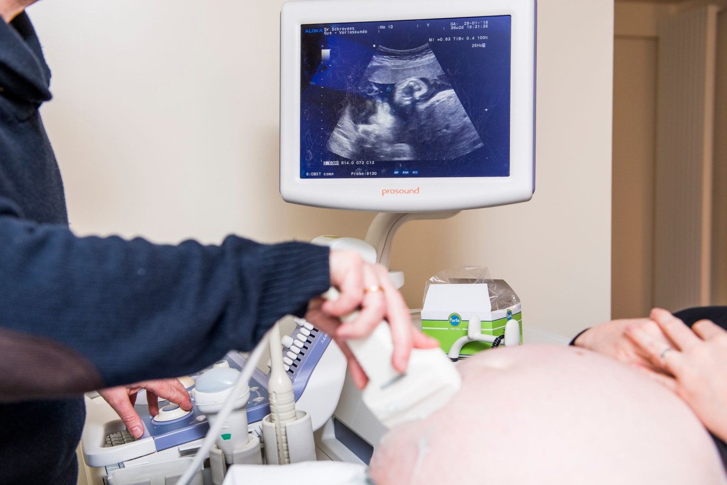 Ein Arzt untersucht eine schwangere Frau mit einem Ultraschallgerät während der Fötus im Mutterbauch auf einem Display zu sehen ist.