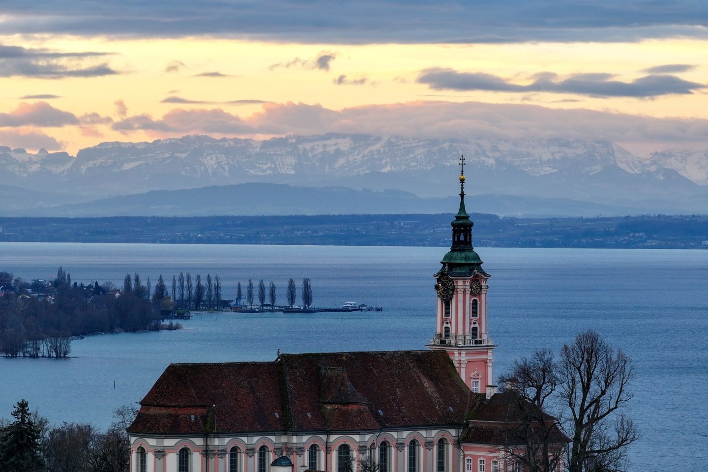 Hinter der ehemaligen Klosterkirche Birnau am Bodensee geht die Sonne auf. Die Alpen sind im Hintergrund zu sehen.