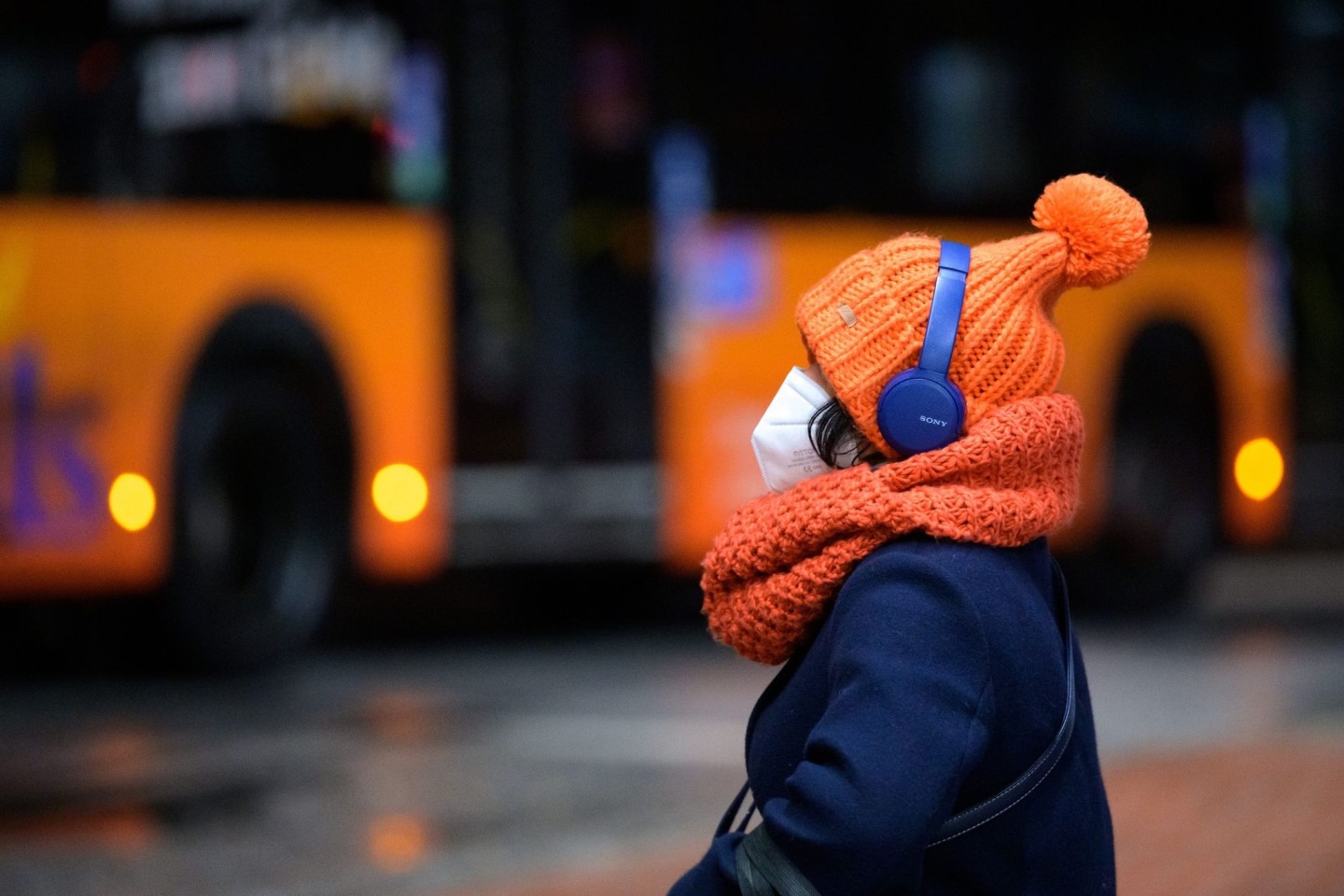 Eine Frau sitzt mit FFP2-Maske, dicker Mütze und Kopfhörern an einem Busbahnhof in Hamburg.