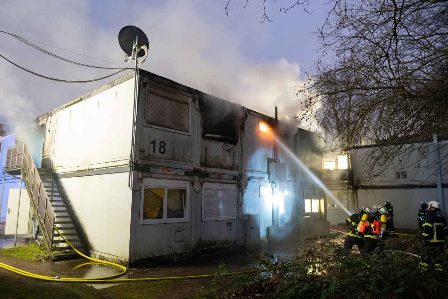 Feuerwehrleute stehen bei Löscharbeiten vor den verqualmten und teils brennenden Container-Einheiten einer Flüchtlingsunterkunft in Hamburg-Winterhude.