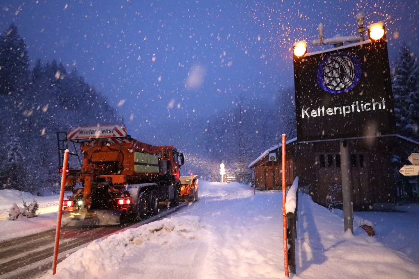 Ein Schild weist auf die Schneekettenpflicht am Riedbergpass im bayerischen Landkreis Oberallgäu hin, während daneben ein Räumfahrzeug den Schnee von der Straße räumt.