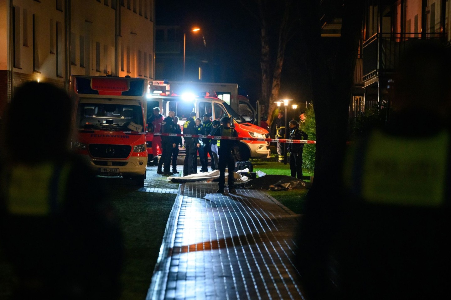 Polizisten, Ermittler und Rettungskräfte stehen am Tatort neben zwei abgedeckten Leichen.